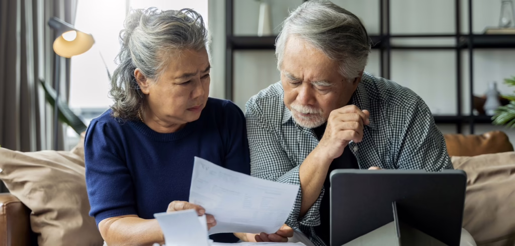 old retired asian senior couple checking and calculate financial billing together on sofa involved in financial paperwork, paying taxes online using e-banking laptop at living room home background
